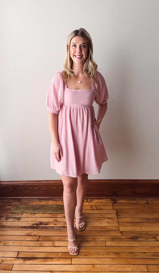 Woman wearing a pink dress standing on a wooden floor with a white wall background