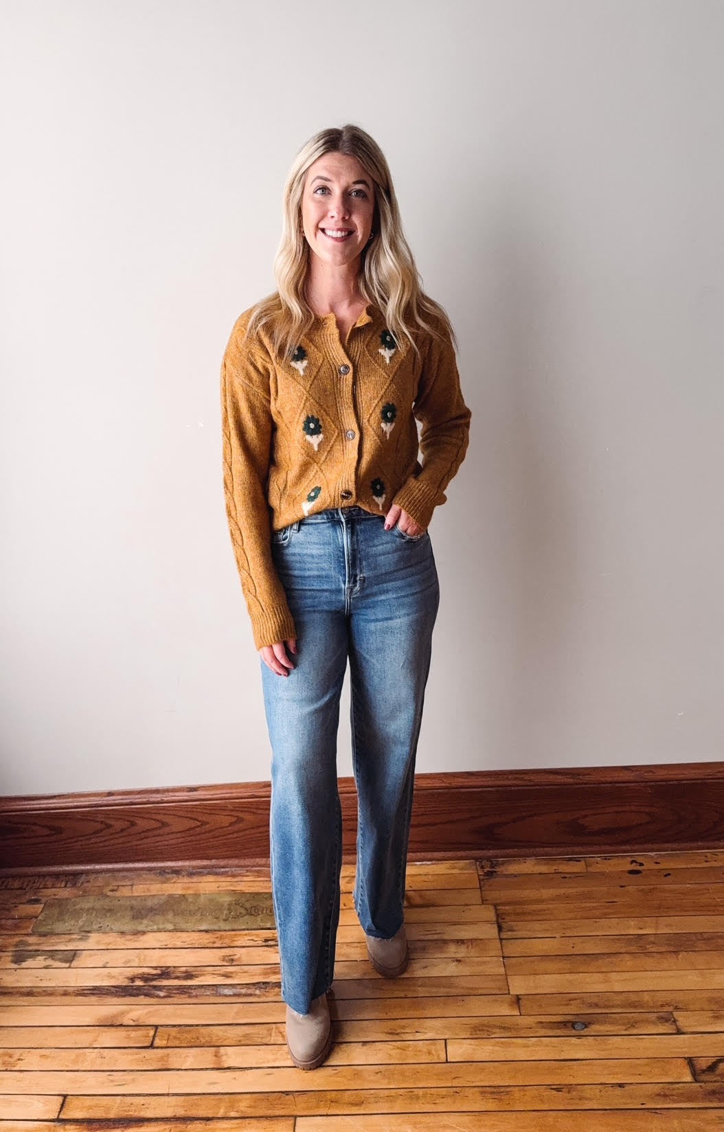 Woman wearing a mustard yellow cardigan and blue jeans standing on a wooden floor.