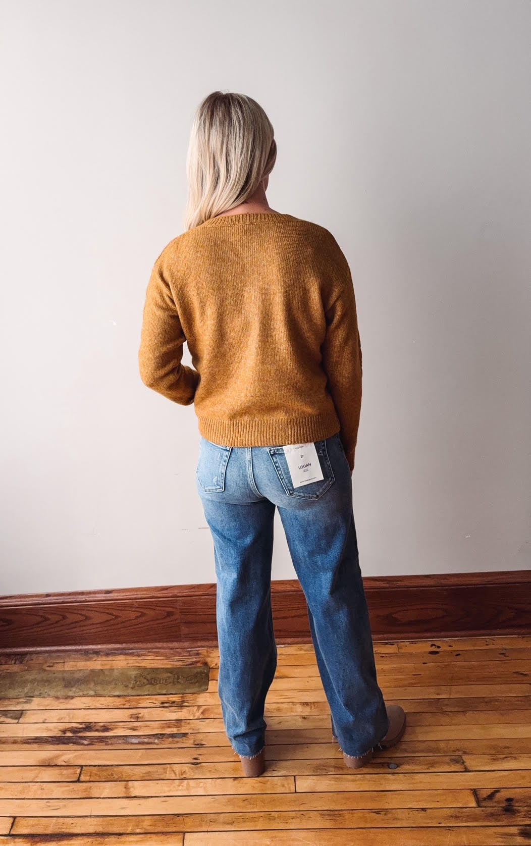 Person wearing a mustard yellow sweater and blue jeans standing in a room with wooden flooring and a white wall.