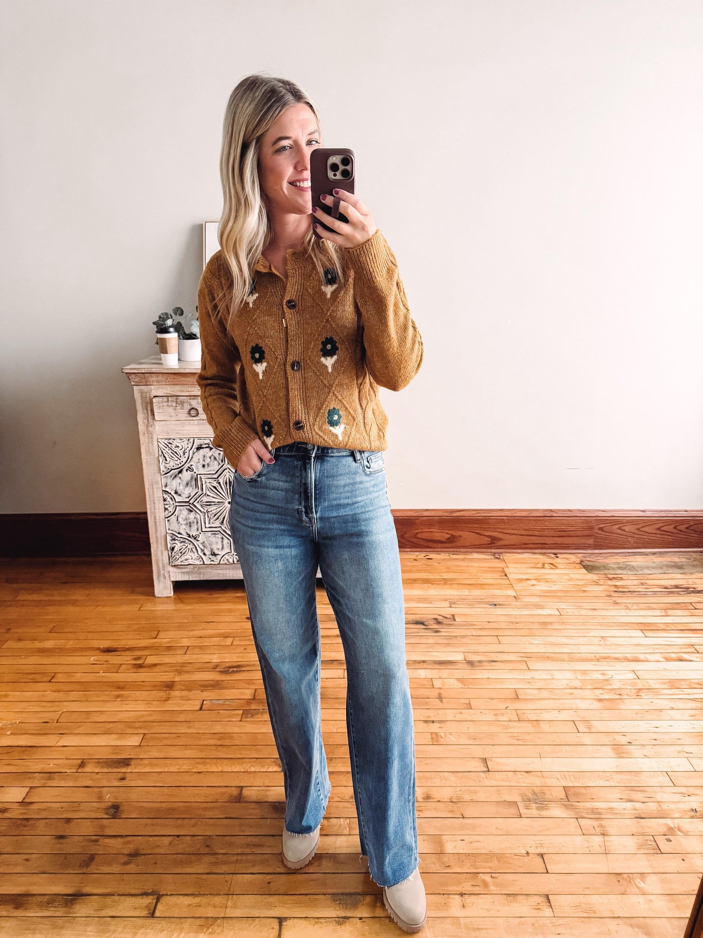 Woman taking a mirror selfie wearing a brown cardigan with colorful buttons and blue jeans.