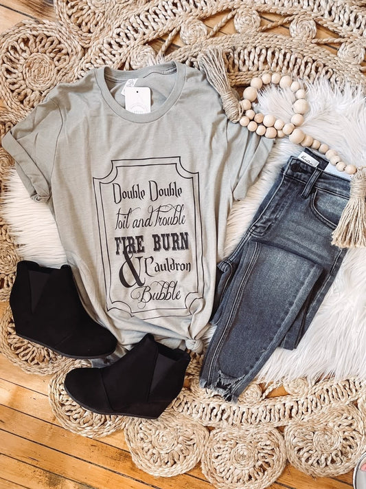 A grey Bella Canvas shirt with the phrase 'Double Double Toil and Trouble' printed on the front, displayed alongside a pair of blue jeans and black ankle boots.