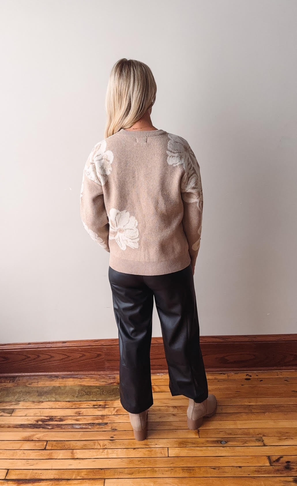 Person wearing a beige sweater with floral patterns and black pants standing on a wooden floor.