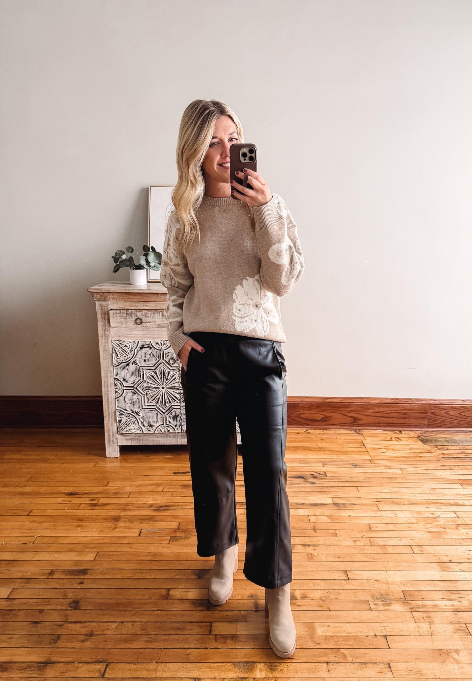 Woman taking a mirror selfie wearing a beige sweater with lace details and black pants in a room with wooden flooring.