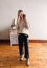 Woman taking a mirror selfie wearing a beige sweater with lace details and black pants in a room with wooden flooring.