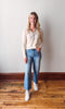 Woman wearing a beige blouse, blue jeans, and white sneakers standing on a wooden floor against a plain wall.