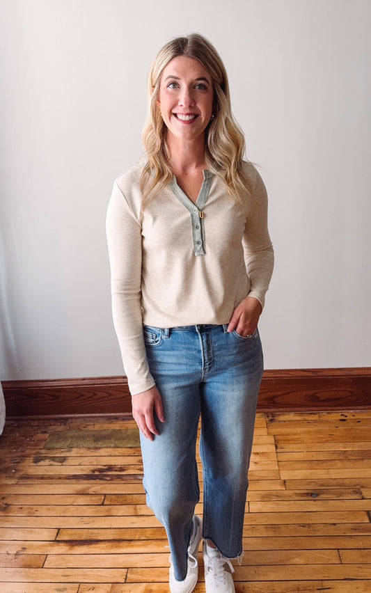 Woman wearing a beige long-sleeve shirt and blue jeans standing on a wooden floor.