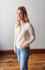 Woman wearing a cream sweater and blue jeans standing against a plain wall.