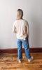 Person wearing a beige sweater and blue jeans standing on a wooden floor against a white wall.