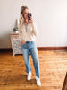 Woman taking a mirror selfie wearing a beige sweater, blue jeans, and white sneakers in a room with wooden flooring.