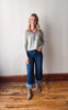 Woman wearing a gray sweater and blue jeans standing on a wooden floor with a white wall background