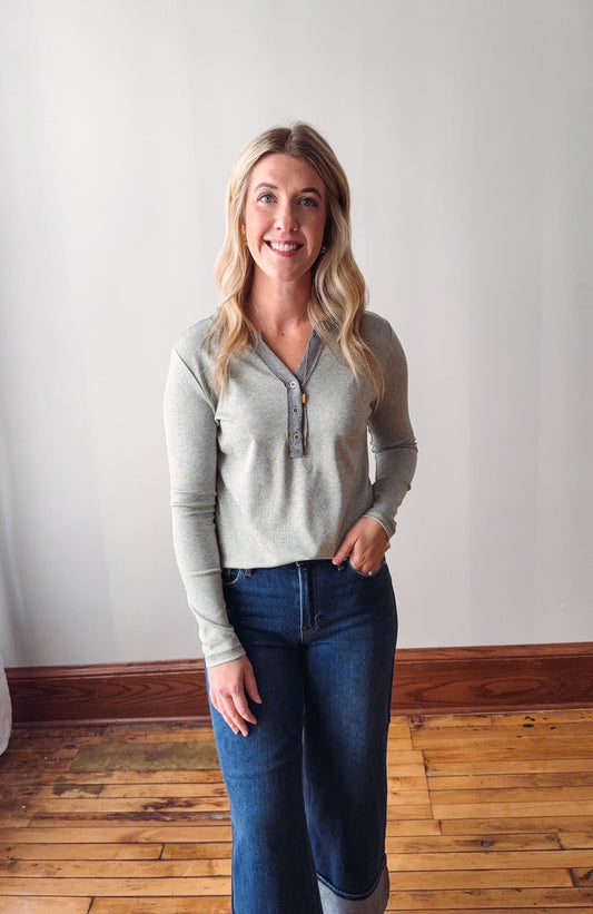 Woman wearing a light gray long-sleeve shirt and blue jeans standing against a white wall.