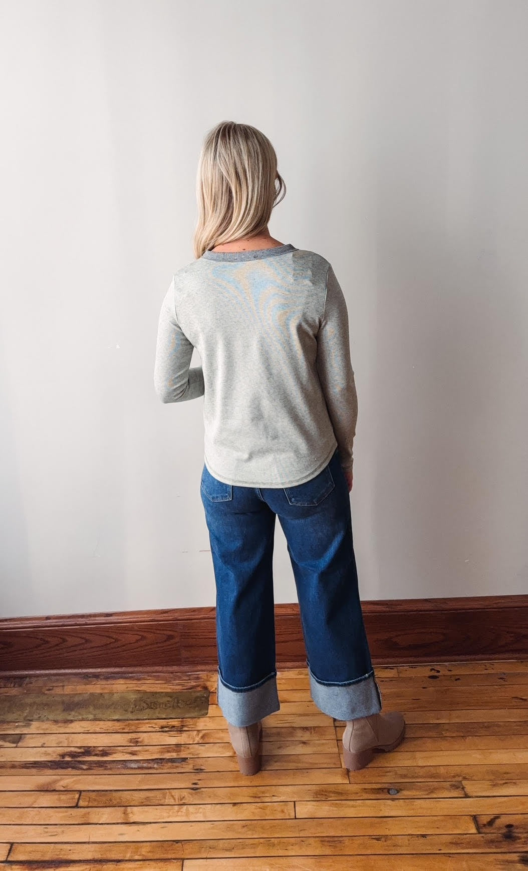 Person wearing a gray sweater and blue jeans standing on a wooden floor against a white wall.