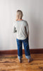 Person wearing a gray sweater and blue jeans standing on a wooden floor against a white wall.