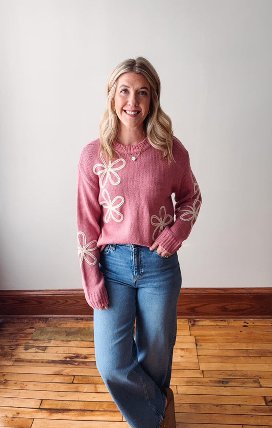 Woman wearing a pink sweater with floral designs standing on a wooden floor.