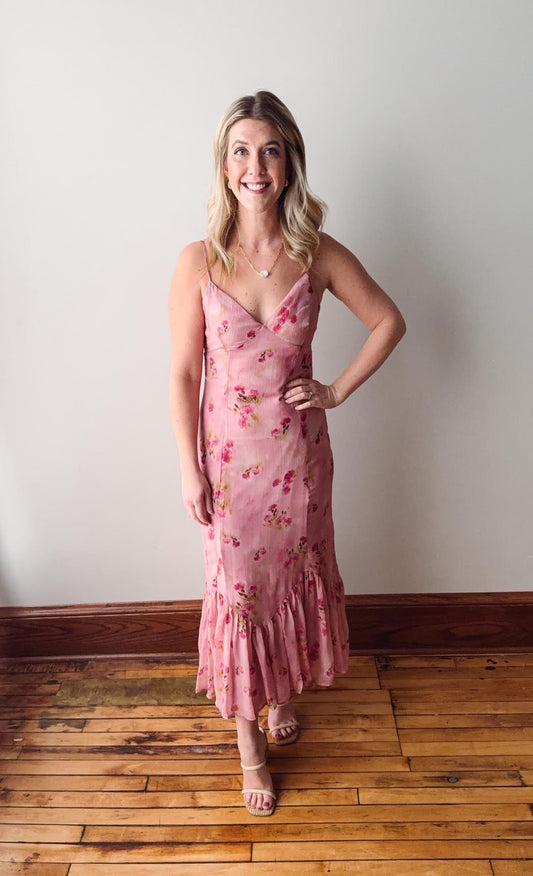 Woman wearing a pink floral dress standing against a white wall on a wooden floor.
