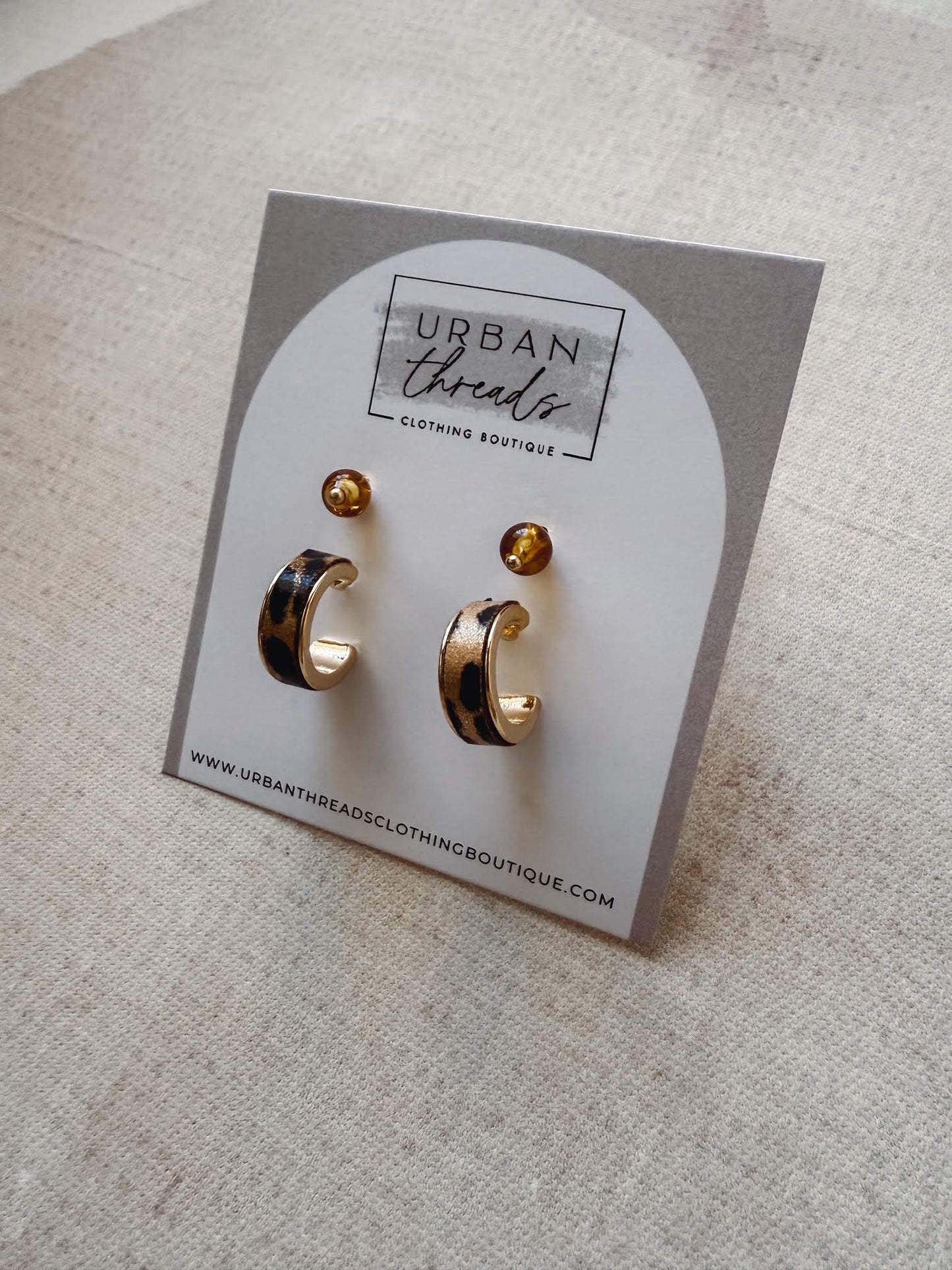 Gold earrings on a card with 'Urban Threads' branding on a beige surface