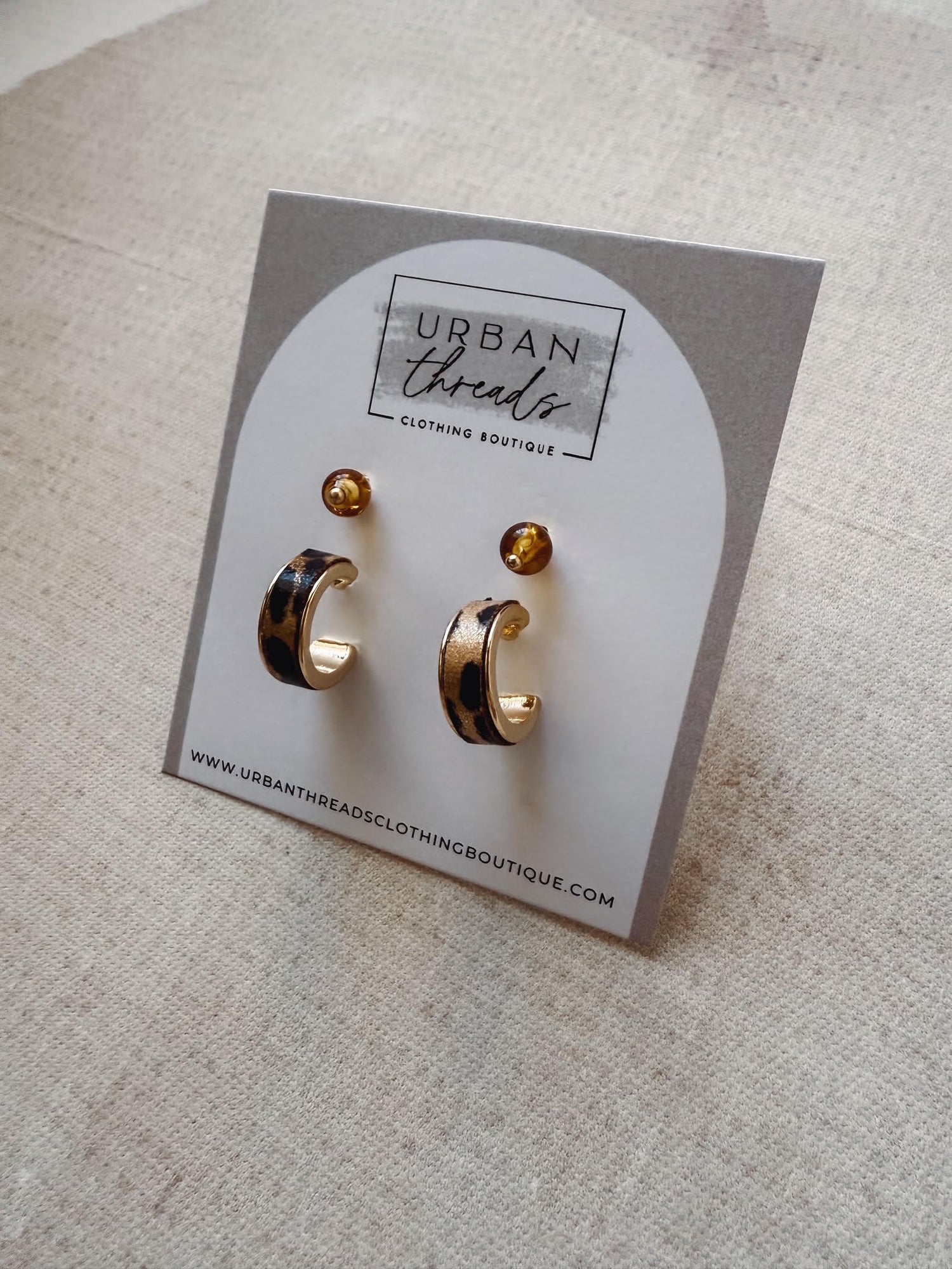 Gold earrings on a card with 'Urban Threads' branding on a beige surface