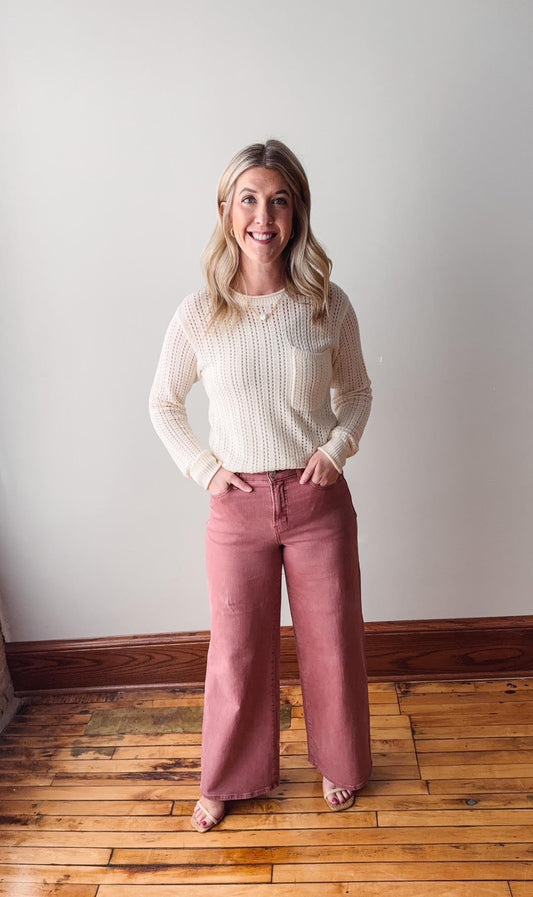 Woman wearing a white sweater and pink pants standing on a wooden floor against a plain wall.
