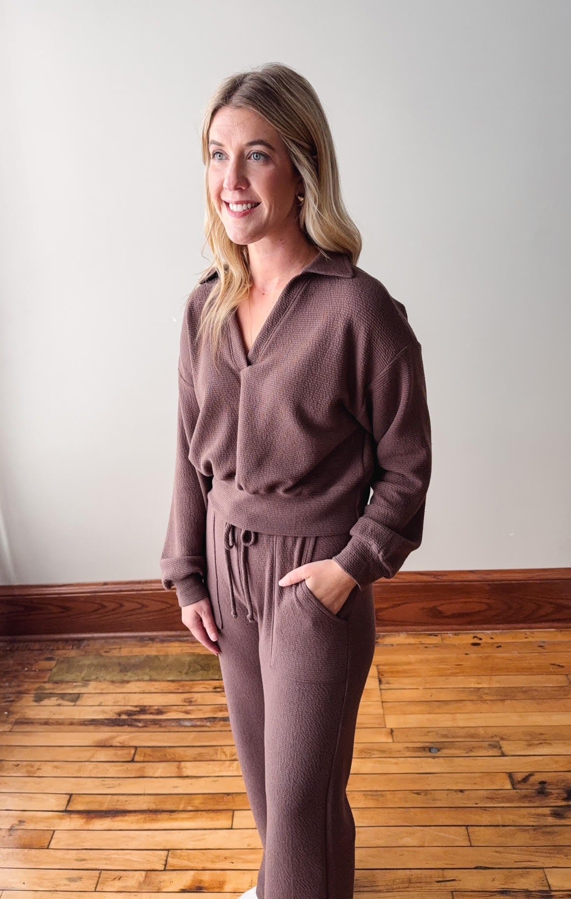 Woman wearing a brown outfit standing on a wooden floor with a white wall background