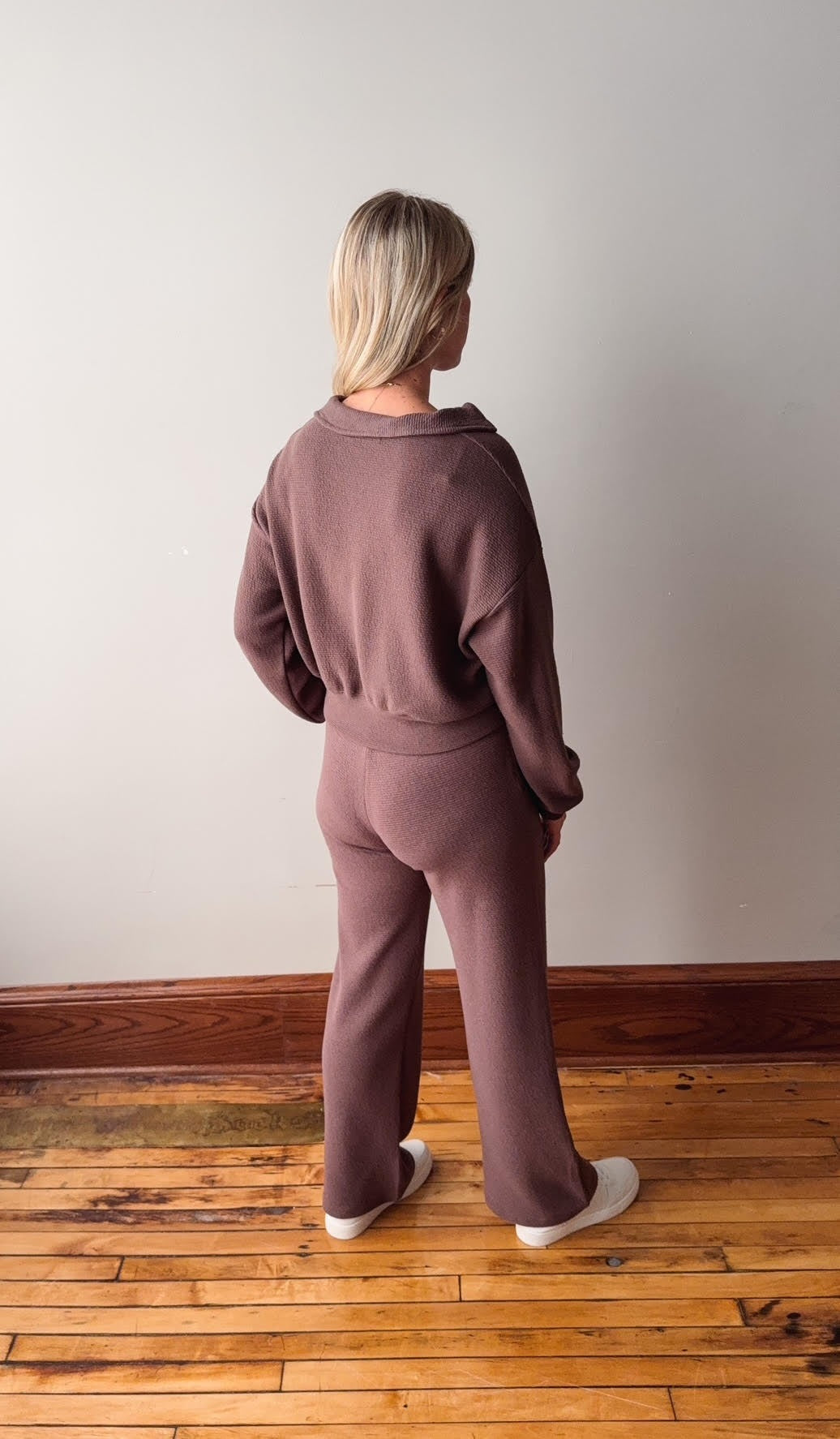 Person wearing a mauve tracksuit standing in a room with wooden flooring and a white wall.