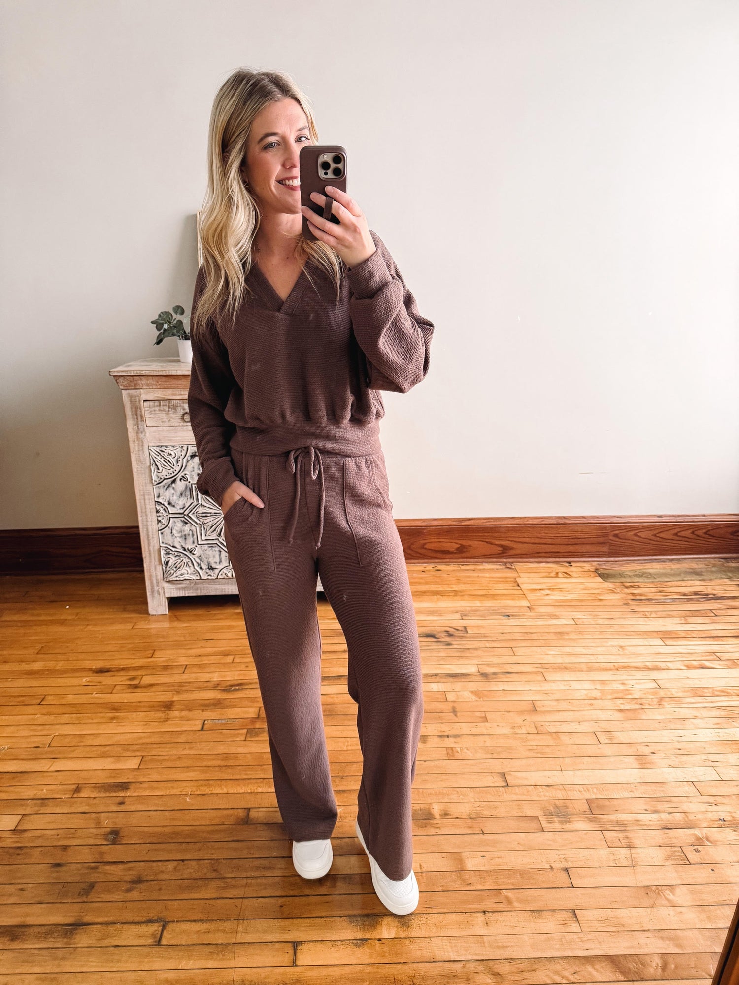 Woman taking a mirror selfie wearing a brown tracksuit in a room with wooden flooring.