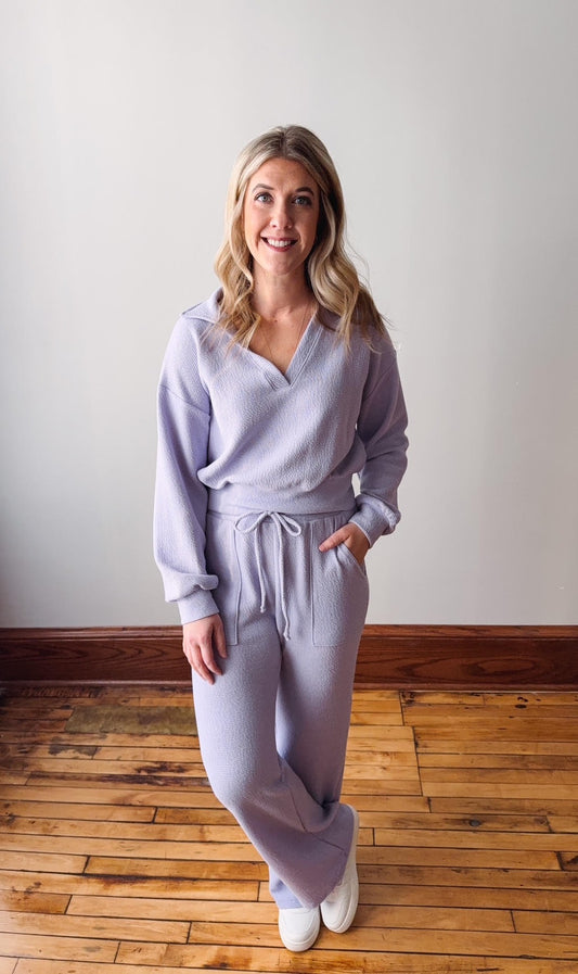 Woman wearing a light purple outfit standing on a wooden floor with a white wall background