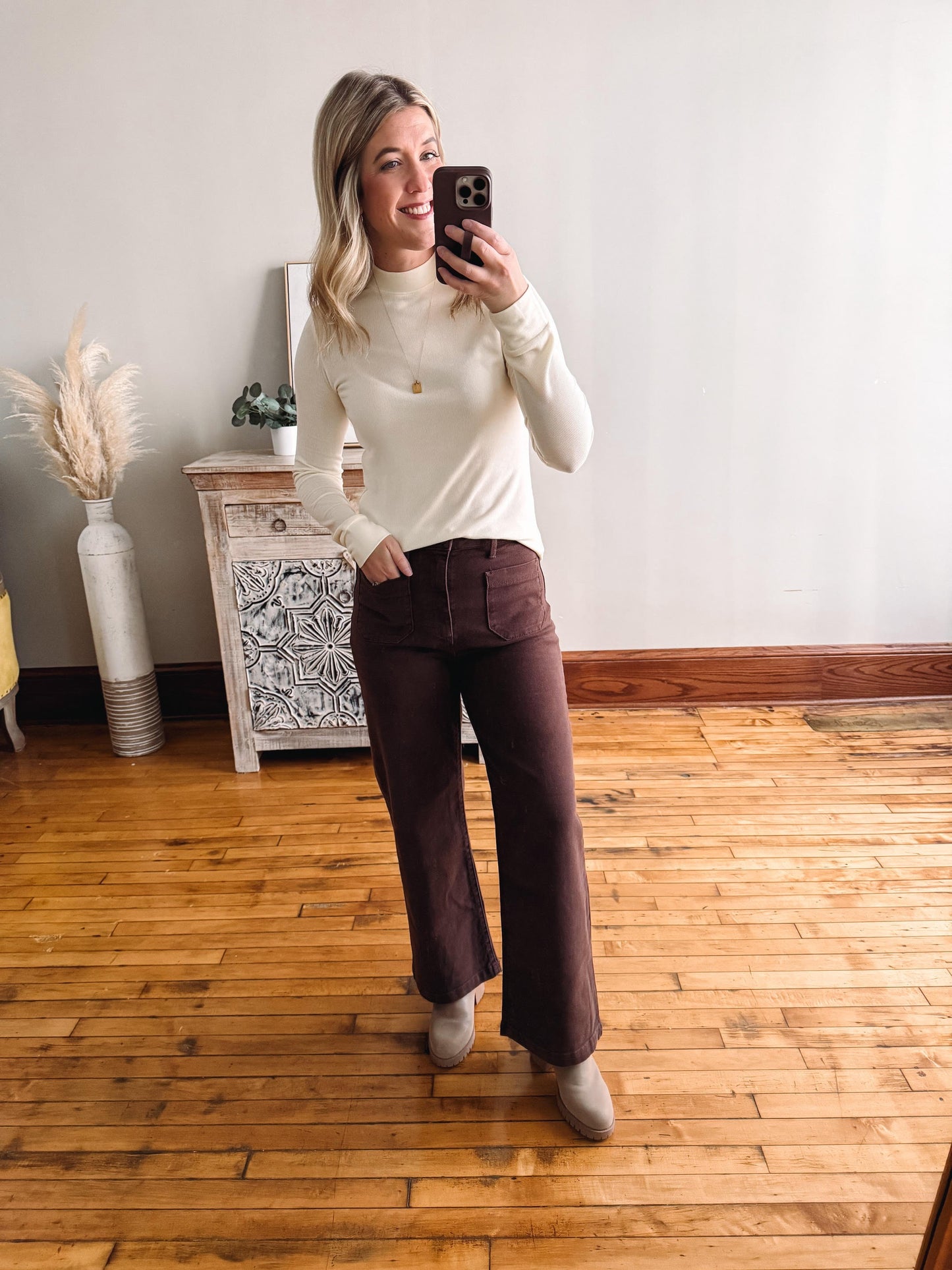 Woman taking a mirror selfie wearing a white turtleneck and brown pants in a room with wooden flooring.
