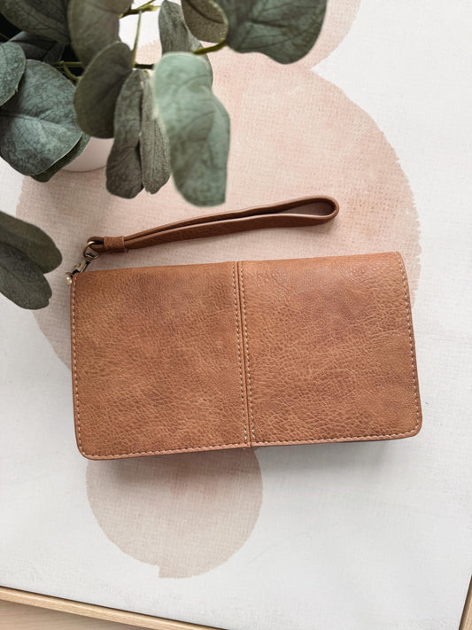 Brown leather wallet with wrist strap on a light background with green leaves.