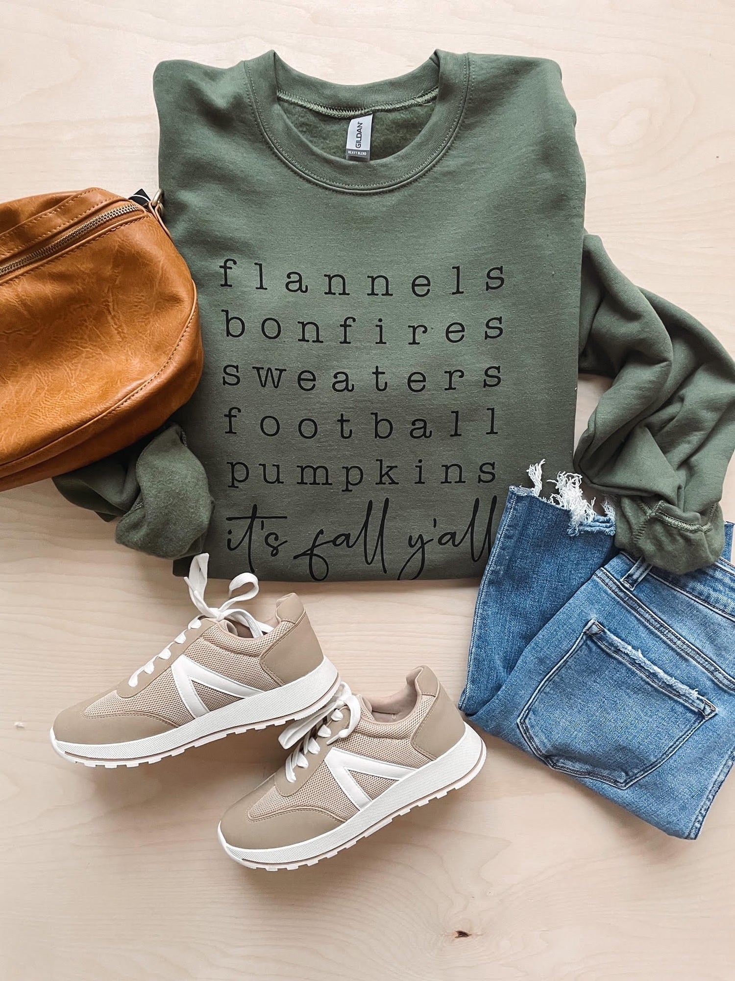 Flat lay of olive green sweatshirt with black text, pair of blue denim jeans, brown leather handbag, and beige sneakers.