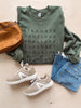 Flat lay of olive green sweatshirt with black text, pair of blue denim jeans, brown leather handbag, and beige sneakers.