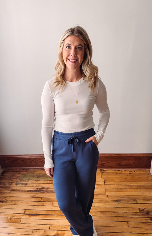 Woman wearing a white long-sleeve top and blue pants standing on a wooden floor.