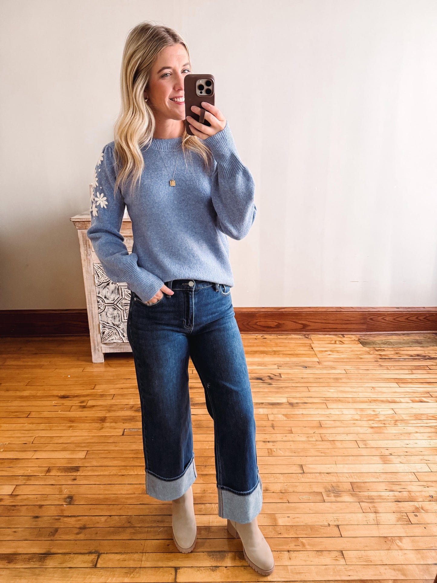 Woman taking a mirror selfie wearing a blue sweater and jeans on a wooden floor.