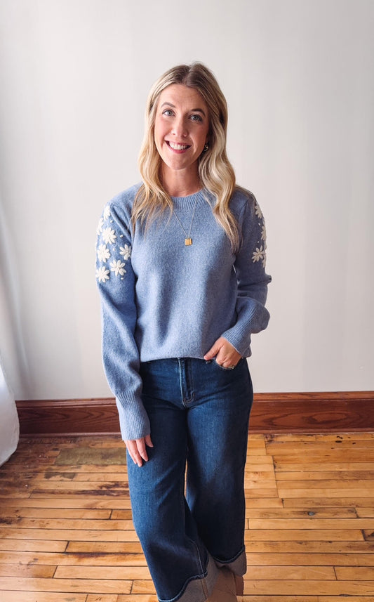 Woman wearing a blue sweater with white patterns and jeans standing on a wooden floor.