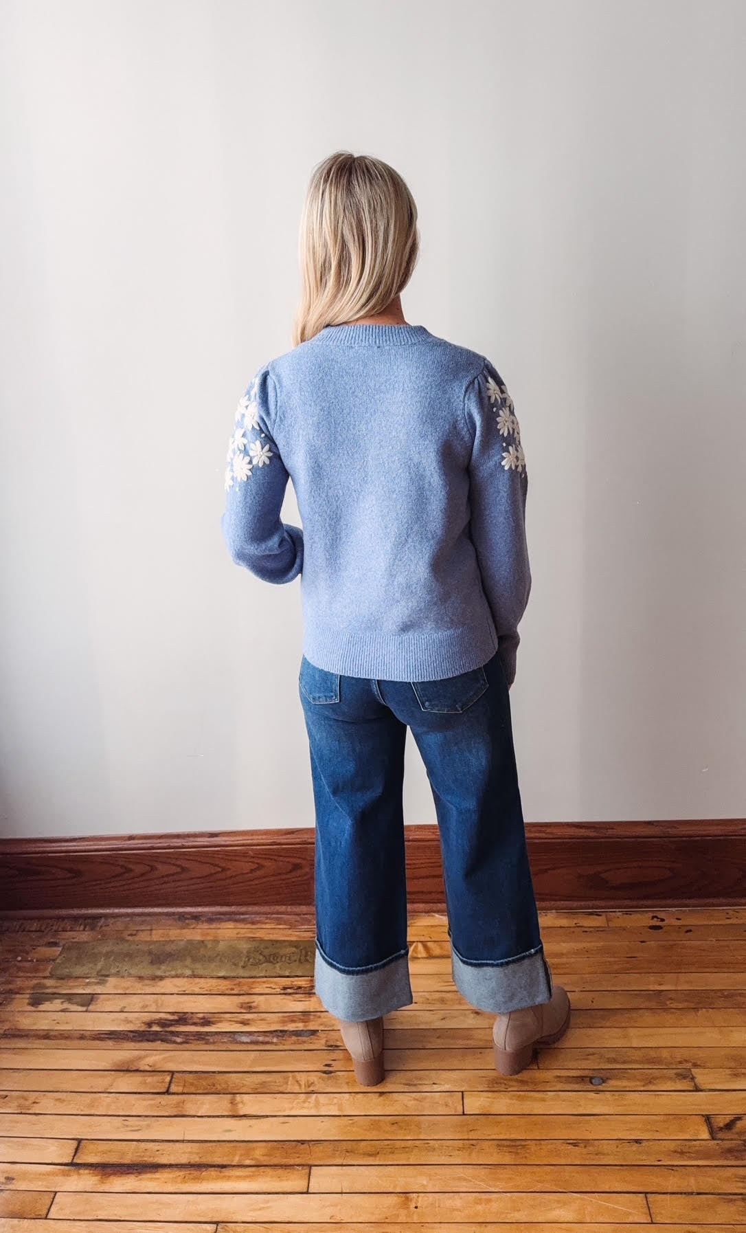 Person wearing a light blue cardigan with floral patterns and dark blue jeans standing on a wooden floor.