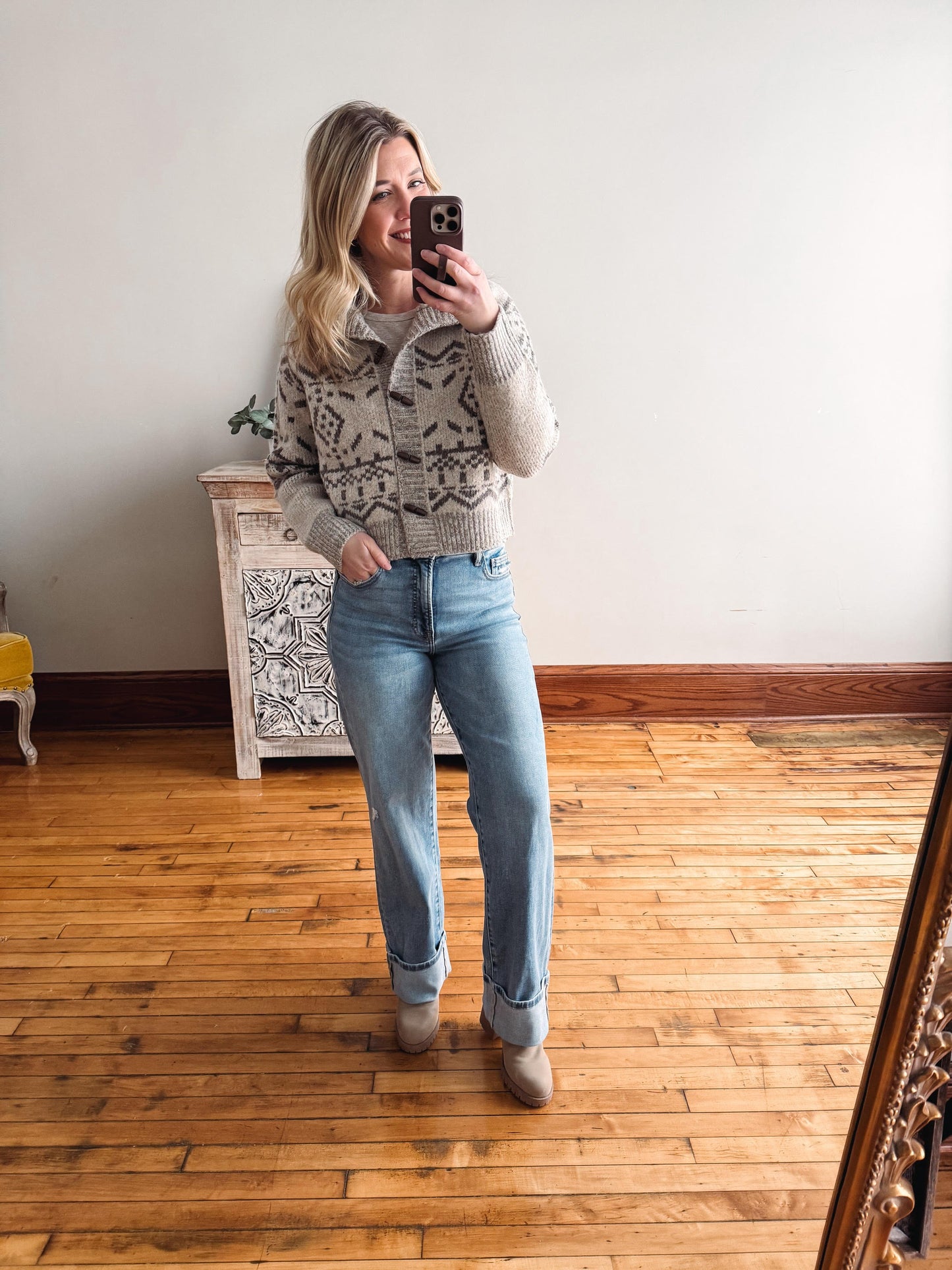 Woman taking a mirror selfie wearing a patterned sweater and jeans in a room with wooden flooring.