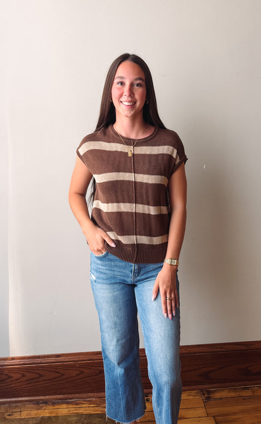 A young woman stands indoors wearing a brown and beige horizontally striped short-sleeve zip-up knit top with blue jeans.