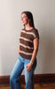 A woman wearing a brown and beige horizontally striped short-sleeve knit top and blue jeans, standing against a plain light gray wall.