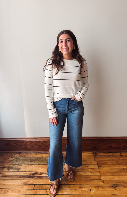 A woman stands in a room with a light beige wall and wooden floor, wearing a cream and black horizontally striped long-sleeve knit sweater, blue wide-leg jeans, and brown leather sandals.
