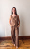 A woman modeling a matching brown linen two-piece set consisting of a sleeveless button-front vest and wide-leg pants, standing in a room with a white wall and wooden floor.