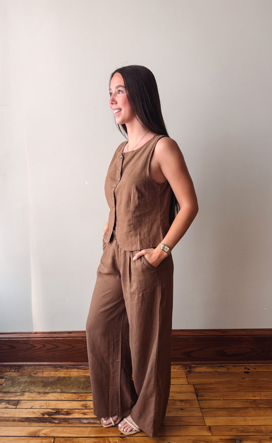 A woman wearing a sleeveless, wide-leg brown jumpsuit with front buttons, standing in profile against a plain off-white wall on a wooden floor.