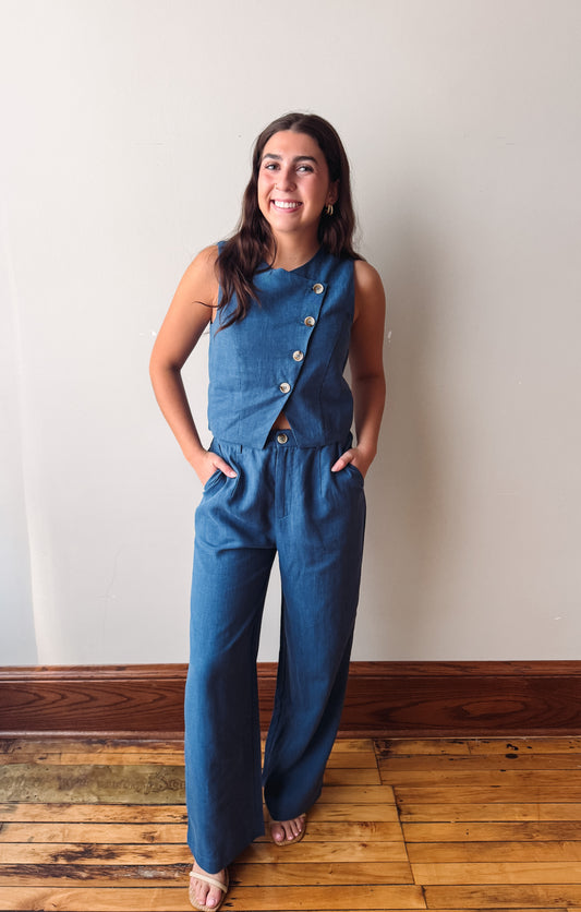A woman modeling a sleeveless blue denim jumpsuit with a buttoned asymmetrical neckline and wide legs, standing on a wooden floor against a plain off-white wall.