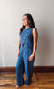 A woman modeling a sleeveless blue wide-leg jumpsuit with decorative buttons down the front, standing on a wooden floor against a plain light gray wall.