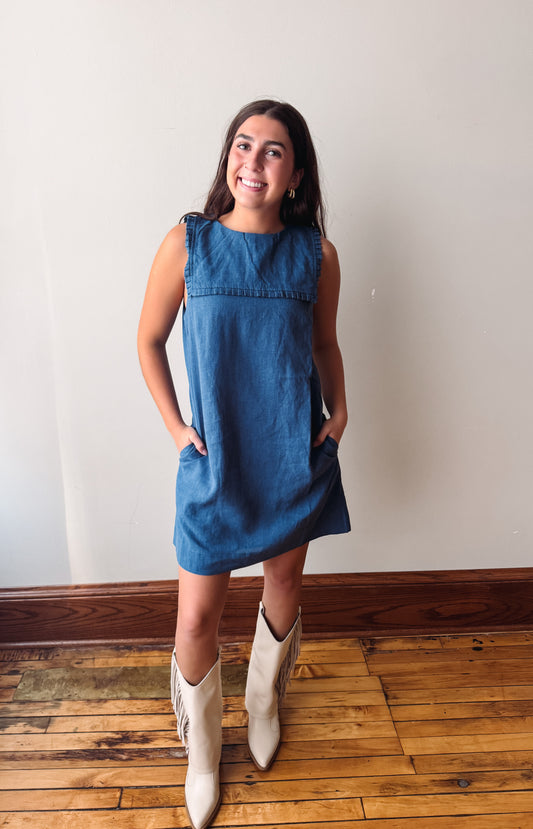 A woman modeling a sleeveless blue denim dress with side pockets, paired with cream-colored cowboy boots, standing on a wooden floor against a plain off-white wall.