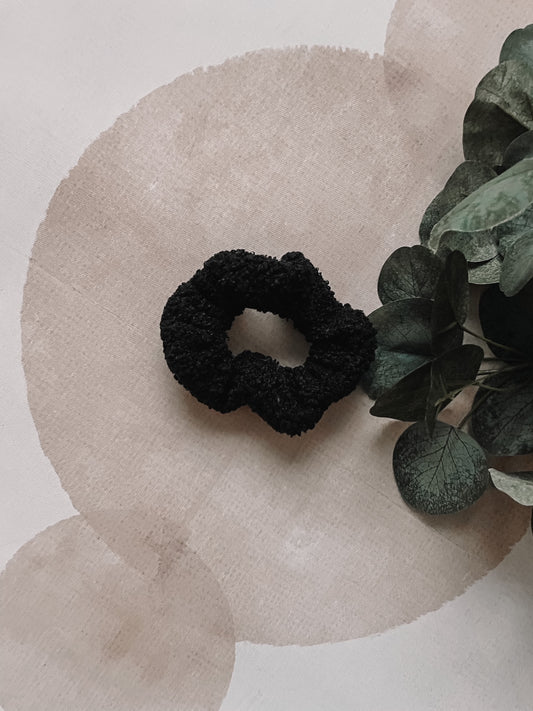 A black sherpa scrunchie placed on a beige background with a plant in the background.