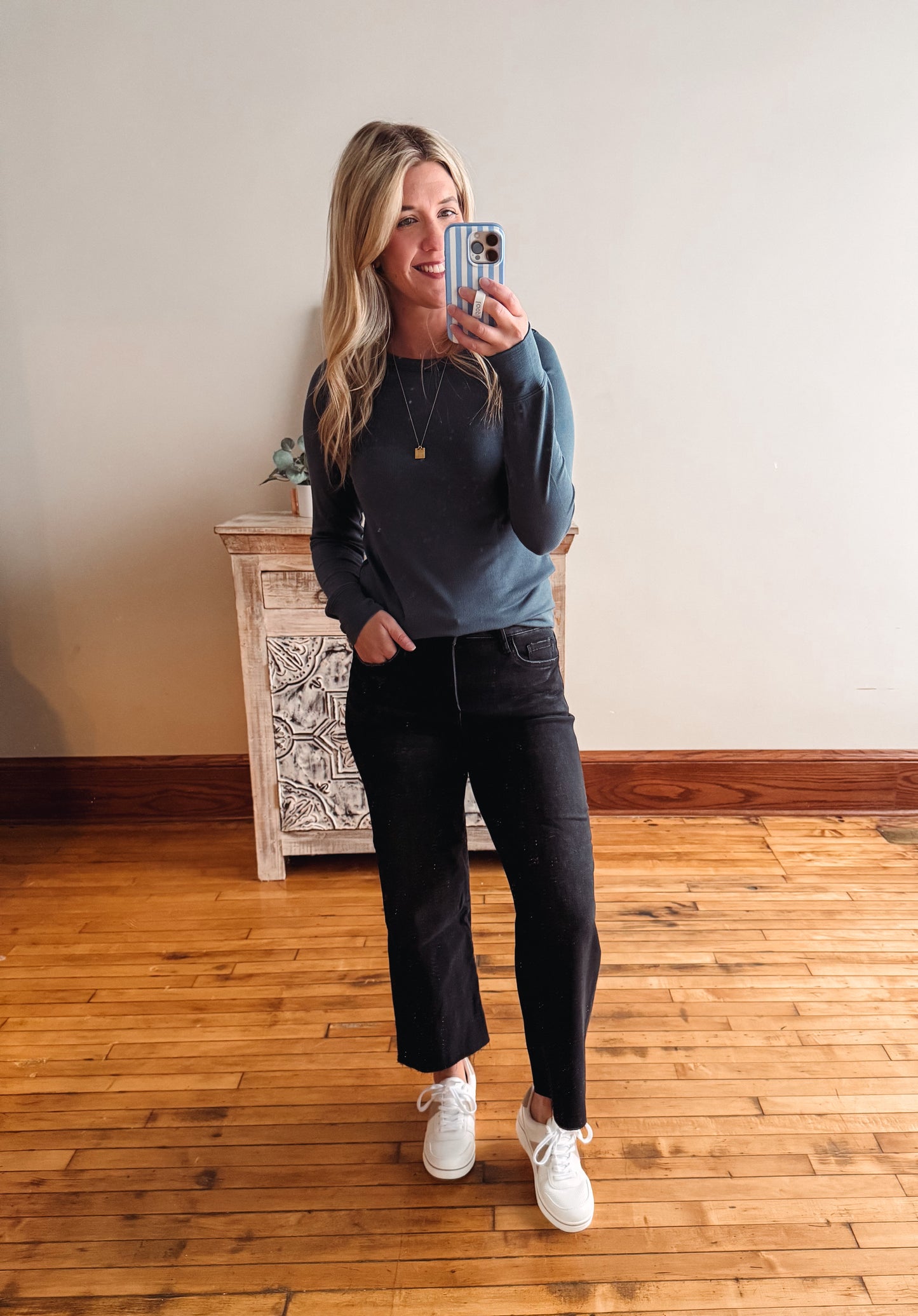 Woman taking a mirror selfie wearing a blue sweater and black jeans on a wooden floor.