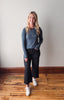 Woman wearing a blue sweater and black pants standing on a wooden floor with a white wall background