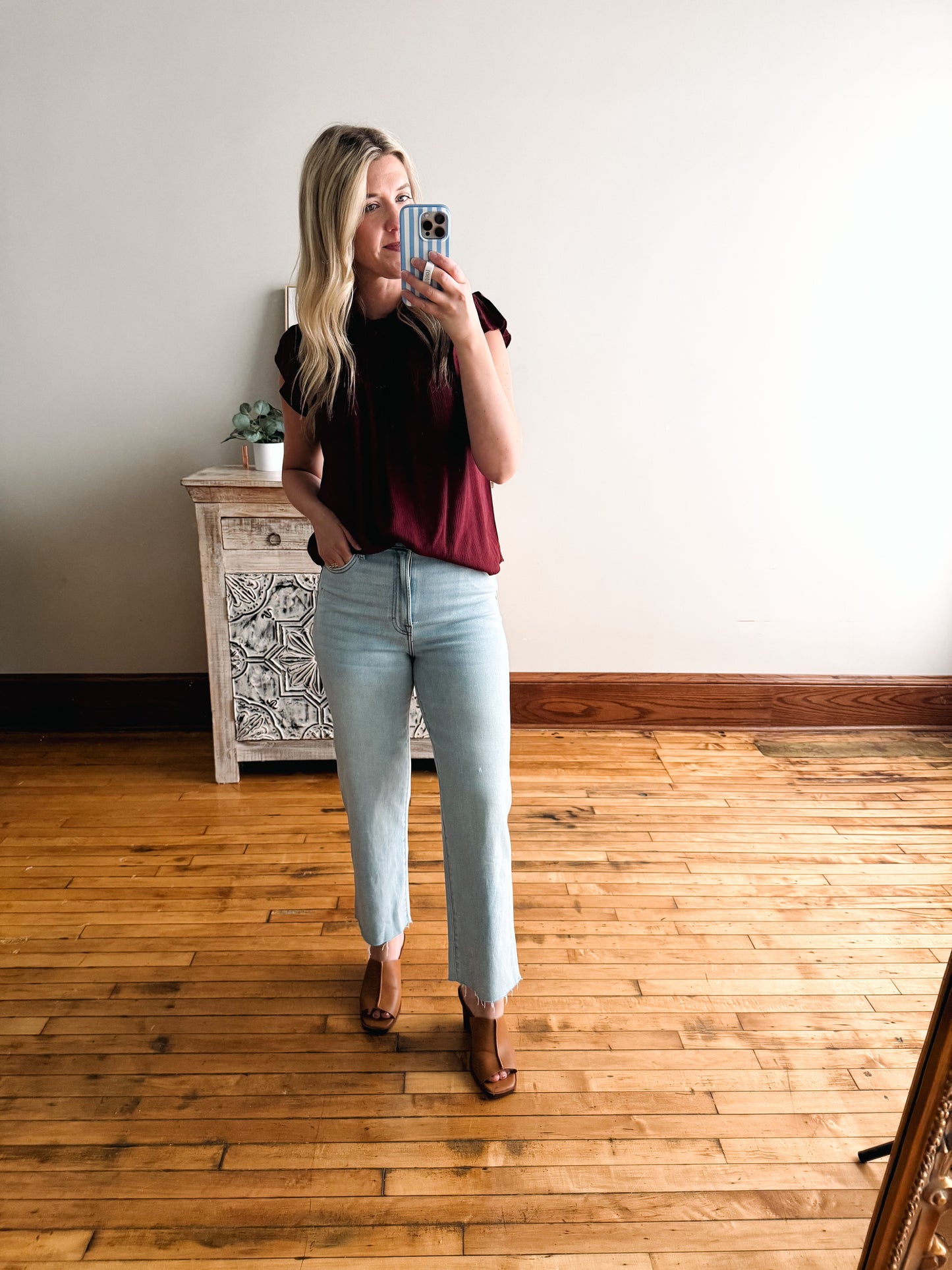 Woman taking a mirror selfie wearing a black top and light blue jeans in a room with wooden flooring.