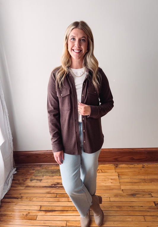 Woman wearing a brown jacket, white shirt, and light blue jeans standing on a wooden floor.