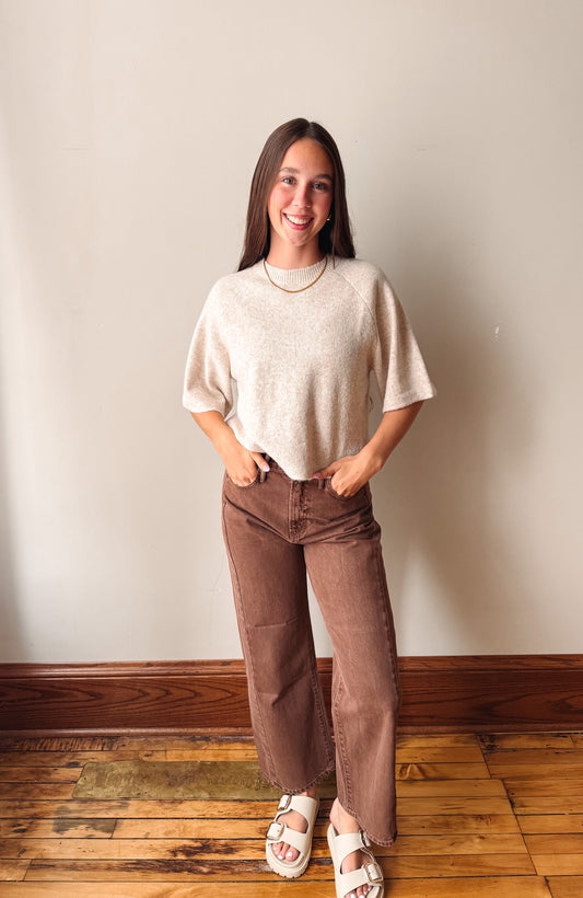 A woman stands indoors wearing a cream-colored short-sleeve knit top, brown wide-leg corduroy pants, and white slide sandals against a plain beige wall and wooden floor.
