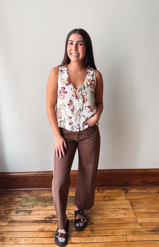 A woman modeling a sleeveless wrap-style sleeveless blouse with a multicolor floral print on a cream background, paired with brown wide-leg pants and black slide sandals, standing in a room with a light gray wall and wooden floor.